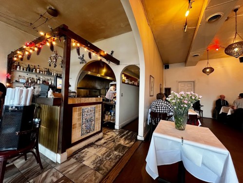 Cozy restaurant interior with arched entry, bar area, and tables set with white linens and flowers. Warm lighting ambiance.
