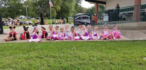A group of young dancers in colorful costumes sitting on a grassy area, with a stage and audience in the background.