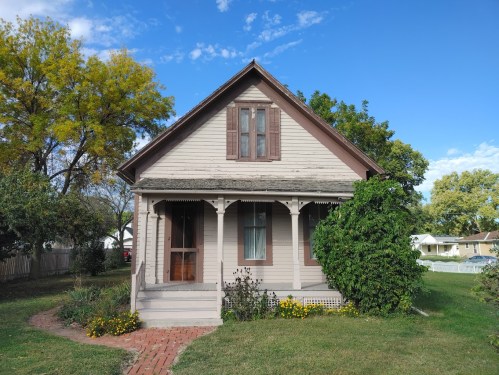 A charming, light-colored house with a front porch, surrounded by greenery and flowers under a blue sky.