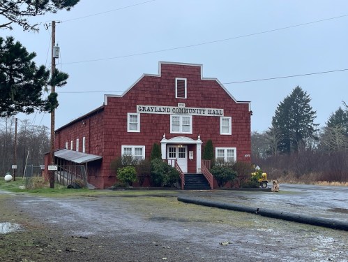 Grayland Community Hall, a red wooden building, surrounded by trees and a gravel area on a cloudy day.