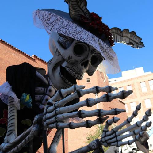 A large skeleton figure wearing a lace hat and floral dress, set against a blue sky and brick buildings.