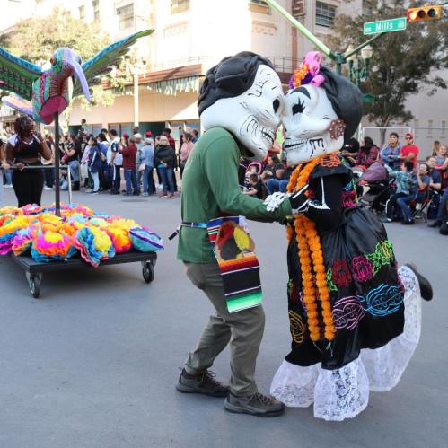 Two dancers in large skull masks perform together in a colorful parade, surrounded by festive decorations and onlookers.