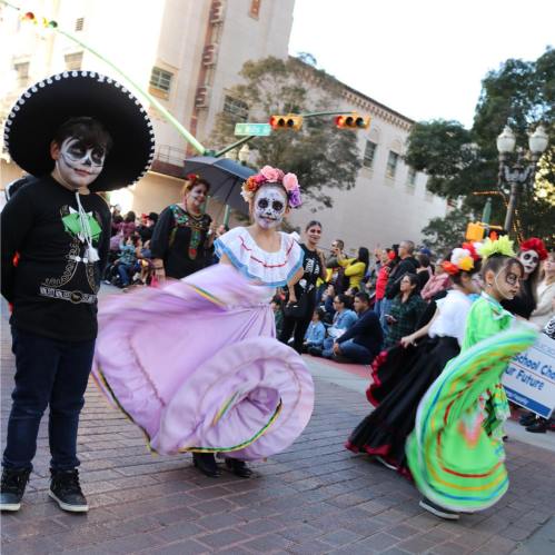 Children in colorful costumes and face paint celebrate at a parade, with a crowd watching in the background.