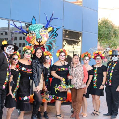 A group of people in colorful costumes and face paint pose together in front of a vibrant, decorated backdrop.