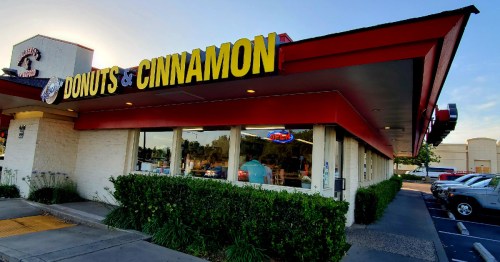 Exterior of a donut shop with a bright sign reading "Donuts & Cinnamon," surrounded by greenery and parked cars.