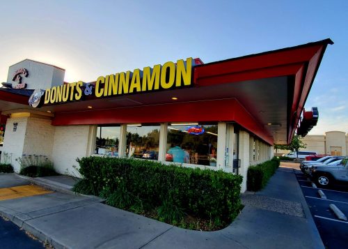 A donut shop with a bright yellow sign reading "Donuts & Cinnamon," surrounded by greenery and parked cars.
