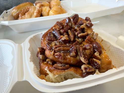 A close-up of a sticky bun topped with pecans, served in a takeout container, with glazed donuts in the background.