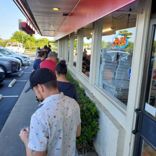 A line of people waiting outside a restaurant with an "OPEN" sign in the window on a sunny day.