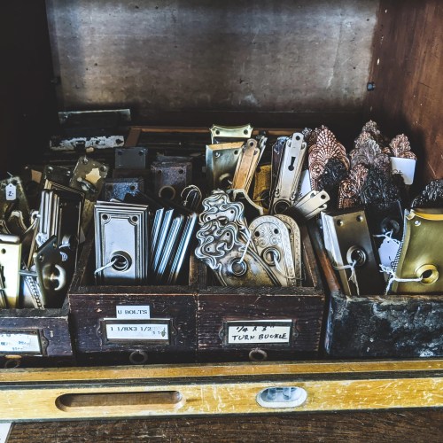A wooden drawer filled with various metal hardware pieces, including bolts, turnbuckles, and decorative fasteners.