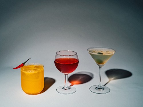 Three colorful cocktails on a gray background: a yellow drink with a chili, a red drink, and a green martini.