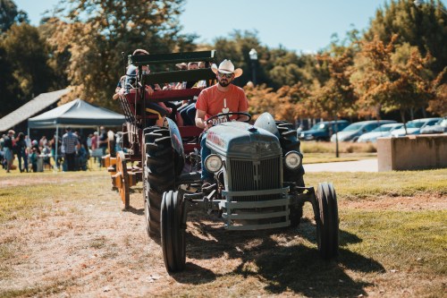 A man in a cowboy hat drives a tractor pulling a wagon with people at an outdoor event. Trees and tents are in the background.