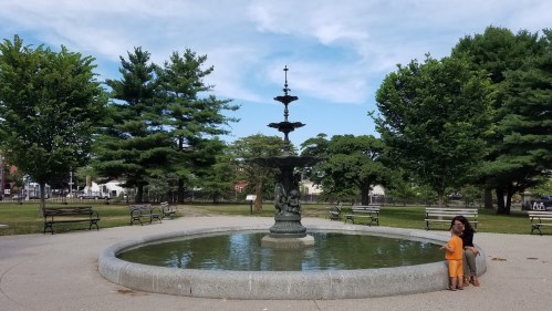 A park scene featuring a fountain surrounded by trees, with two children playing near the water's edge.