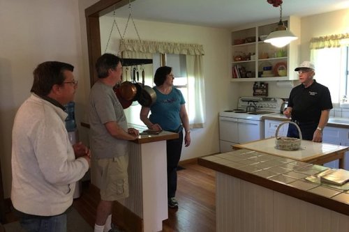 Four people are engaged in conversation in a bright kitchen with white cabinets and hanging pots.