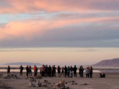 A group of people stands on a beach at sunset, with colorful clouds and mountains in the background.