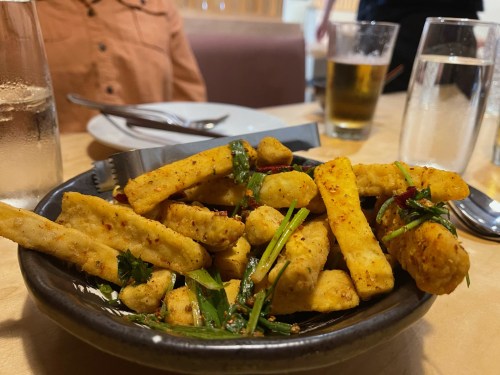 A bowl of crispy fried snacks garnished with herbs and spices, served on a table with drinks in the background.