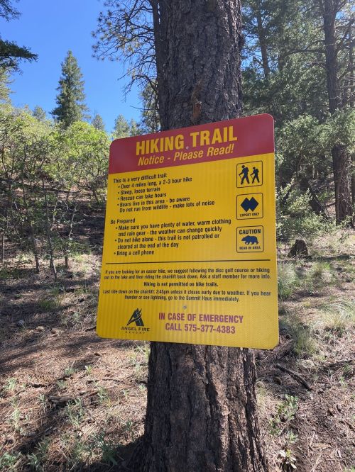 A sign on a tree warns about hiking trail safety, advising water, clothing, and emergency contact information.