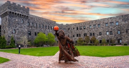 A statute of a wizard holding a red orb stands in the courtyard of a castle-like building beneath a sky pink with sunset.