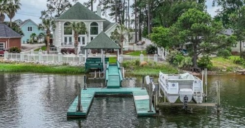 A waterfront property with a green dock, boat lift, and a large house surrounded by trees and a white fence.