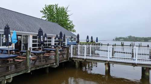 A waterfront restaurant with outdoor seating, umbrellas, and a wooden deck, surrounded by calm water on a cloudy day.