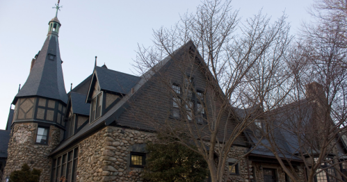 A large, historic stone mansion with a steep roof and multiple gables, surrounded by trees and greenery.