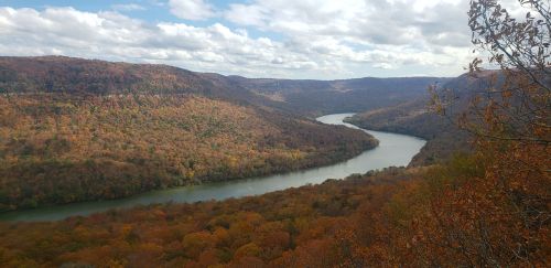 A winding river flows through a valley surrounded by autumn-colored trees under a partly cloudy sky.