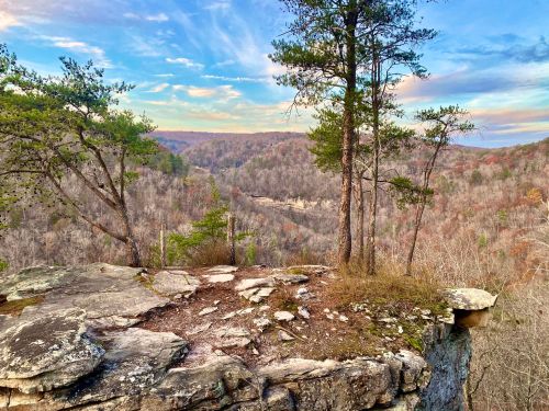 A rocky cliff overlooks a valley with trees and rolling hills under a colorful sky at sunset.