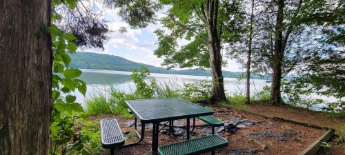A picnic area with a green table and benches by a serene lake, surrounded by trees and hills in the background.