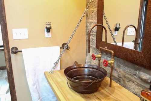 A rustic bathroom sink with a copper basin, vintage-style faucets, and a wooden countertop against a stone wall.