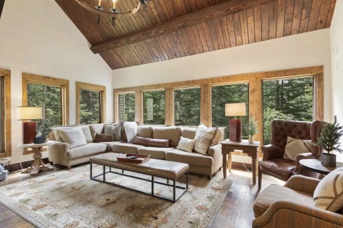 Cozy living room with large windows, wooden beams, a sectional sofa, and a rustic coffee table, surrounded by greenery.