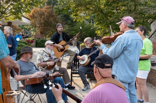 A group of musicians playing instruments outdoors, including guitars, banjos, and a violin, with an audience nearby.