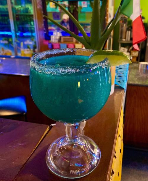 A vibrant blue cocktail in a salt-rimmed glass, garnished with a lime wedge, set against a colorful bar background.
