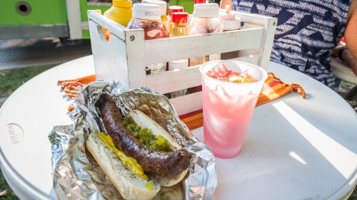 A sausage in a bun with toppings, a drink, and a condiment station on a table outdoors.