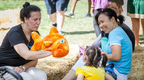 Two women interact with a child, one holding a plush fish puppet, in a sunny outdoor setting with hay bales.