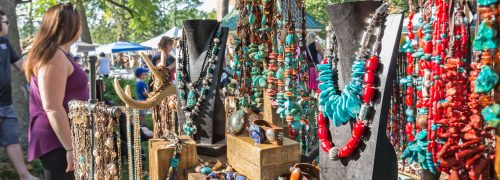 Colorful jewelry displayed on stands at an outdoor market, with people browsing in the background.