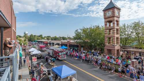 A bustling street parade with crowds, fire trucks, and a clock tower in the background under a blue sky.