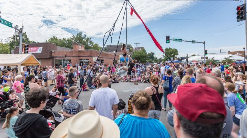 Aerial performer entertains a large crowd at a street festival, with colorful tents and a blue sky in the background.
