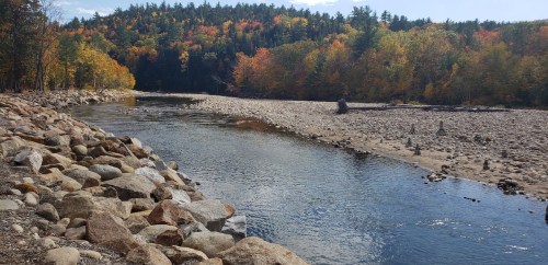 A serene river flows through a rocky landscape, surrounded by trees displaying vibrant autumn colors.