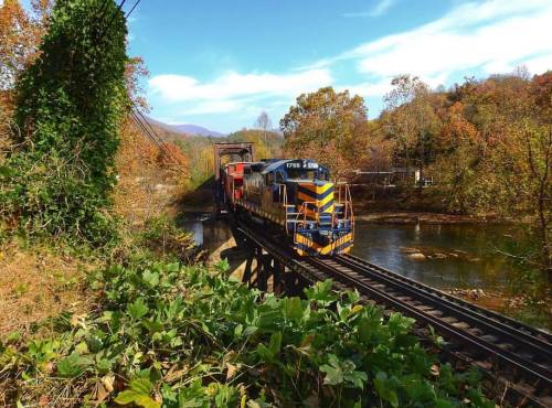 A colorful train crosses a bridge over a river, surrounded by autumn foliage and blue skies.