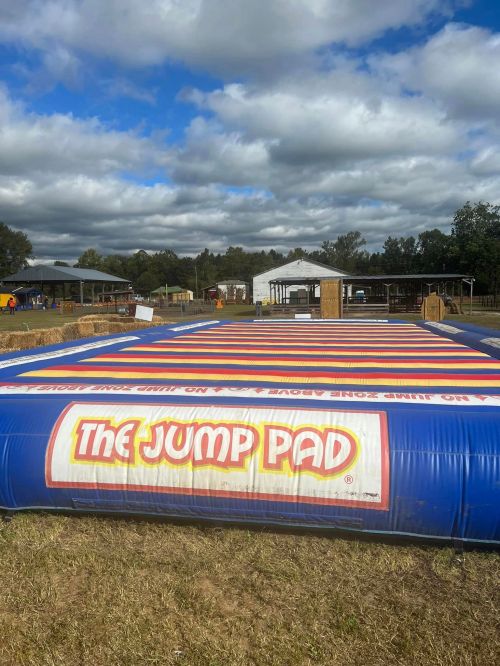 A large inflatable jump pad with "The Jump Pad" logo, set in an outdoor area with cloudy skies and farm structures in the background.