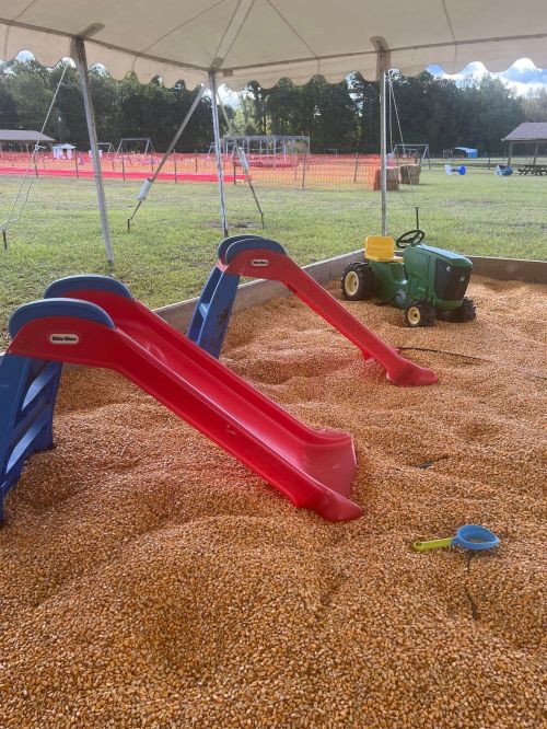 Two small slides in a sandy play area, with a toy tractor and a shovel nearby under a tent.