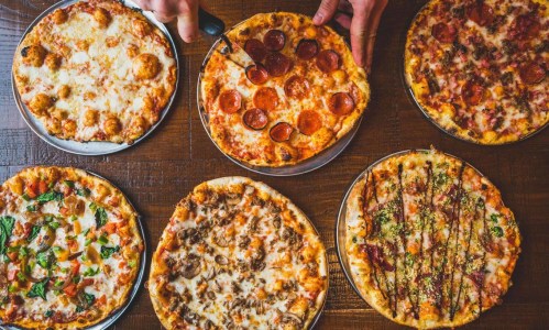 A variety of pizzas on wooden tables, including pepperoni, meat, and veggie options, with hands cutting a slice.