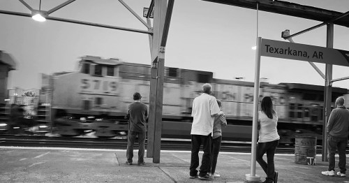 A group of people watches a passing train at Texarkana station, captured in black and white.
