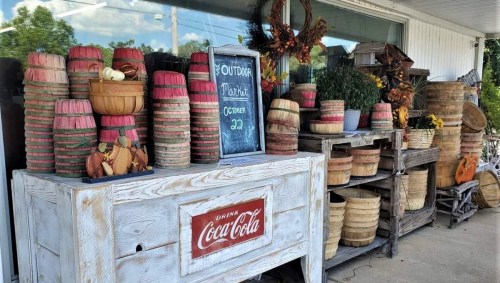 A rustic display of colorful baskets and autumn decorations outside a shop, with a chalkboard sign and Coca-Cola crate.