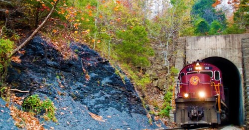 A vintage train emerges from a tunnel surrounded by colorful autumn foliage and rocky terrain.