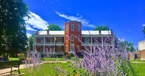 Historic building with a tower, surrounded by purple flowers and green trees under a bright blue sky.