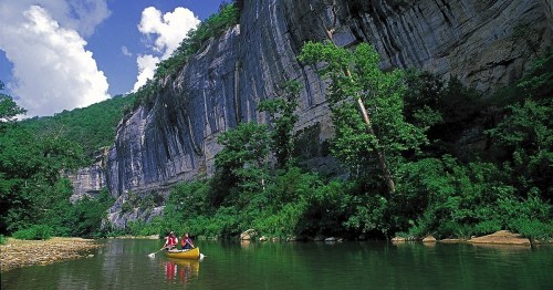 Two people canoeing on a calm river surrounded by lush greenery and towering cliffs under a blue sky with clouds.