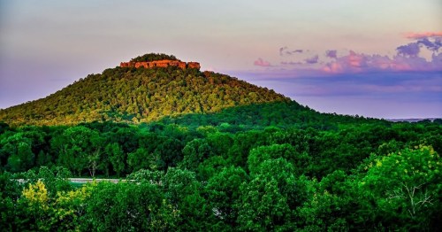 A lush green hill with a rocky summit under a colorful sky, surrounded by dense trees and a winding road below.