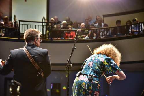 A musician plays on stage while another performs, with an audience watching from a balcony above.