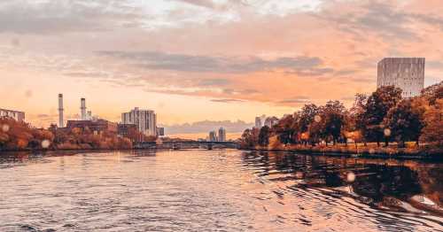 A serene river scene at sunset, with city buildings and trees reflecting on the water's surface.