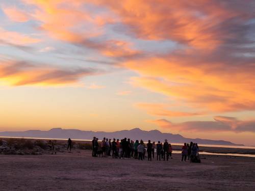 A group of people gathers at sunset, with colorful clouds and mountains in the background.